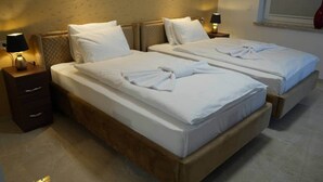 Habitación doble con dos camas, cada una con sábanas y almohadas blancas, cabeceros de madera, mesitas de noche con lámparas y una ventana con persianas.