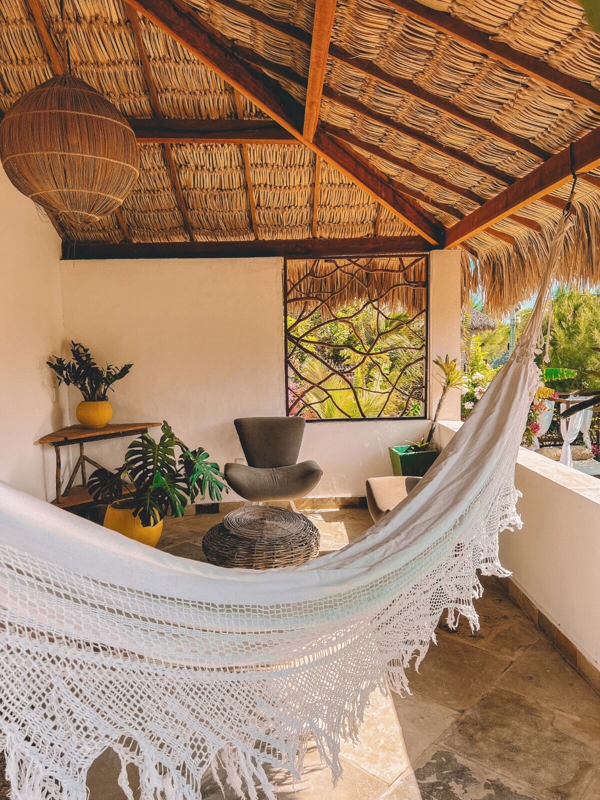 A hammock in a room with a thatched roof, a window with a view of greenery, and several potted plants.