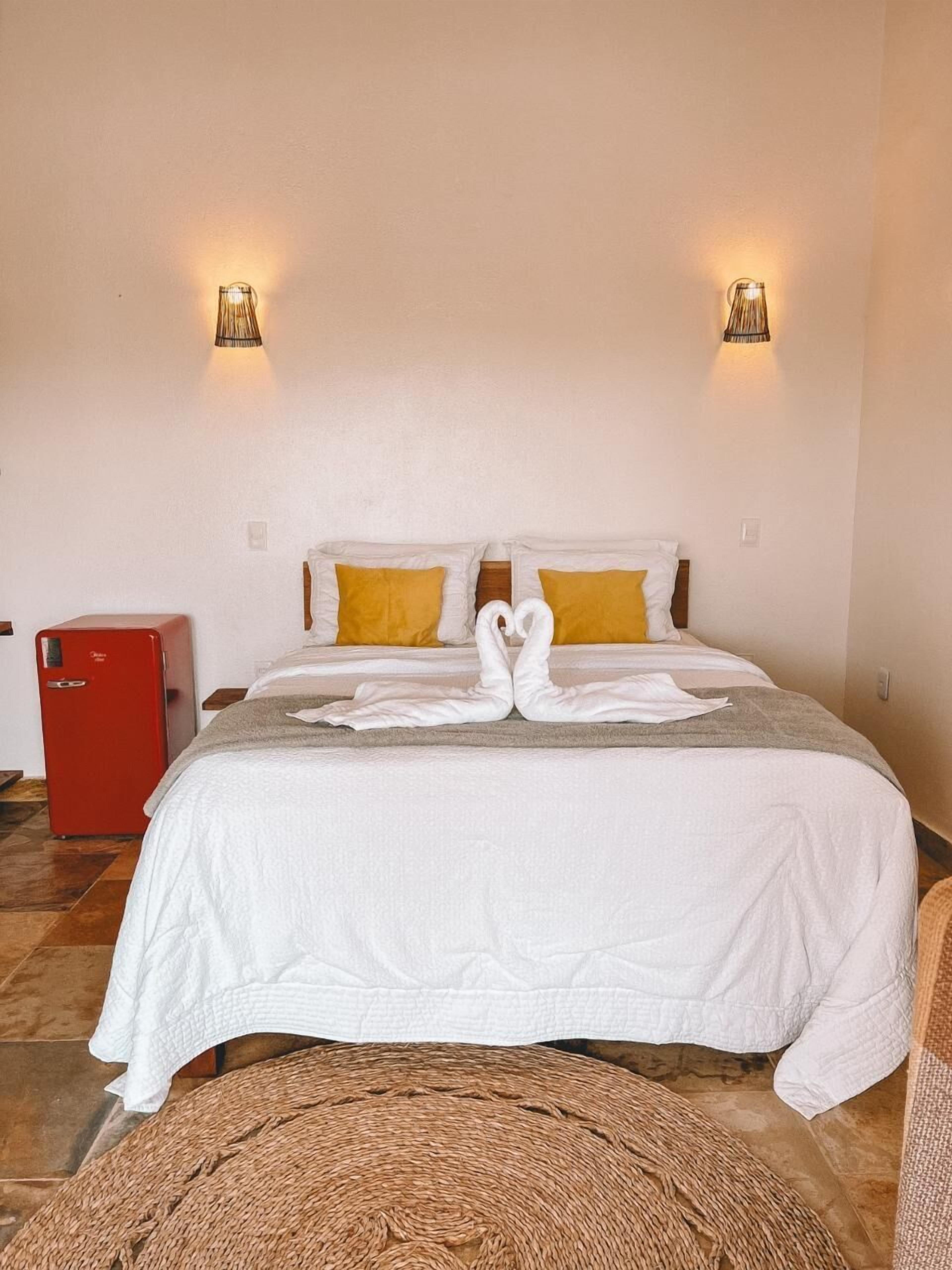 A hotel room with a bed, two wall-mounted lamps, a red refrigerator, and a woven rug.