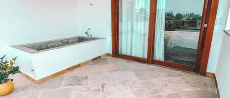 Un balcón con suelo de piedra, una bañera integrada y una puerta de cristal que da a un jardín.