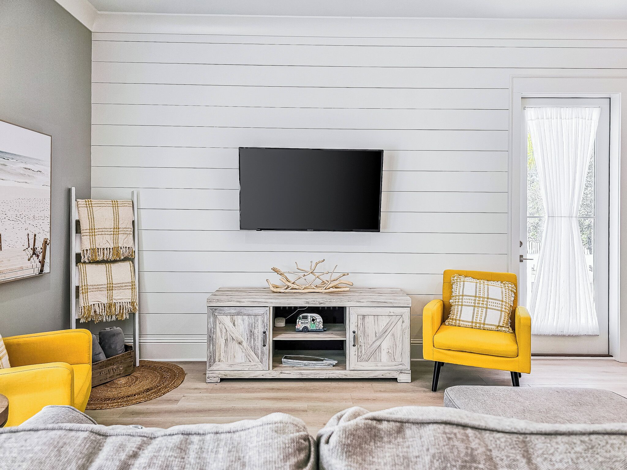 Sala de estar con televisor montado, una mesa de centro de madera con ramas decorativas, un sillón amarillo y un sofá gris.