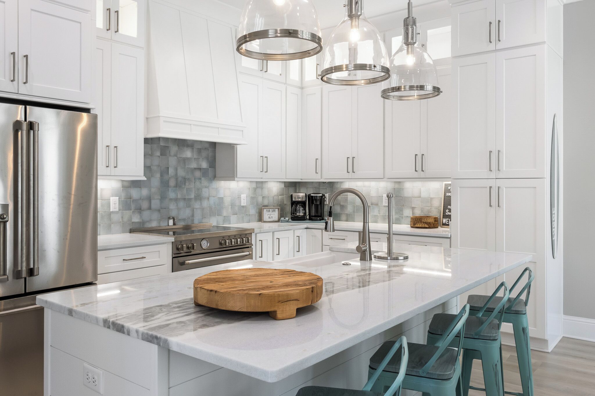 Una cocina moderna con armarios blancos, electrodomésticos de acero inoxidable, encimera de mármol y taburetes de bar color aguamarina.