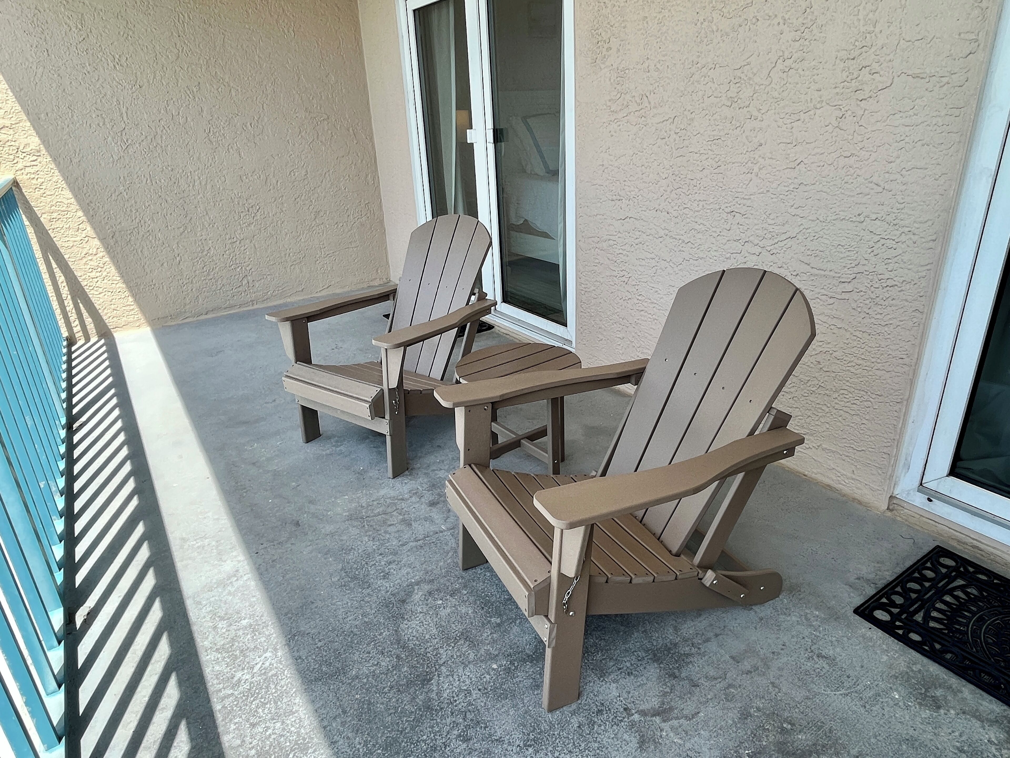 Two Adirondack chairs on a concrete patio with a beige wall and a window in the background.