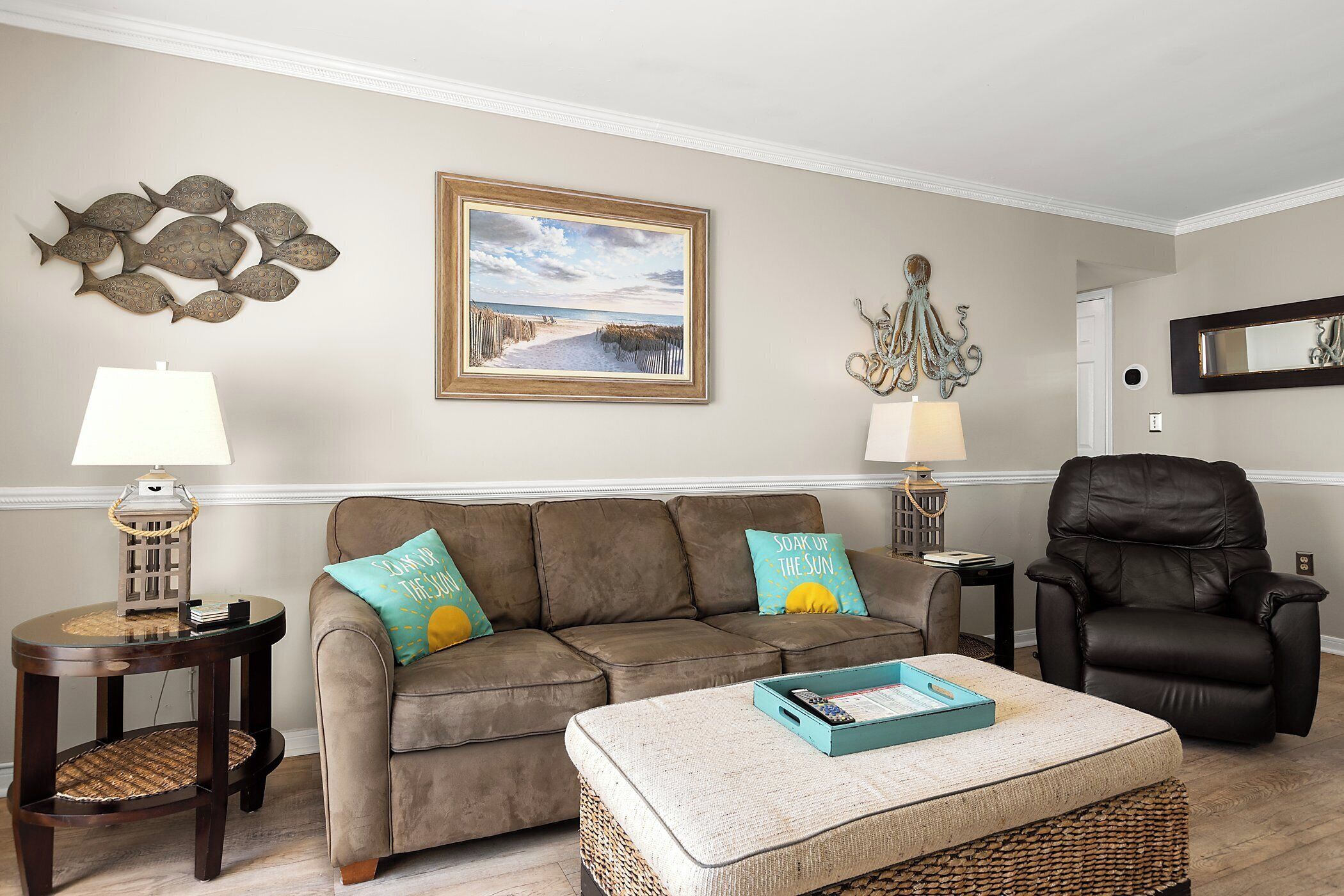 A living room with a brown sofa, two side tables, a black leather chair, a framed beach picture, and decorative wall art.