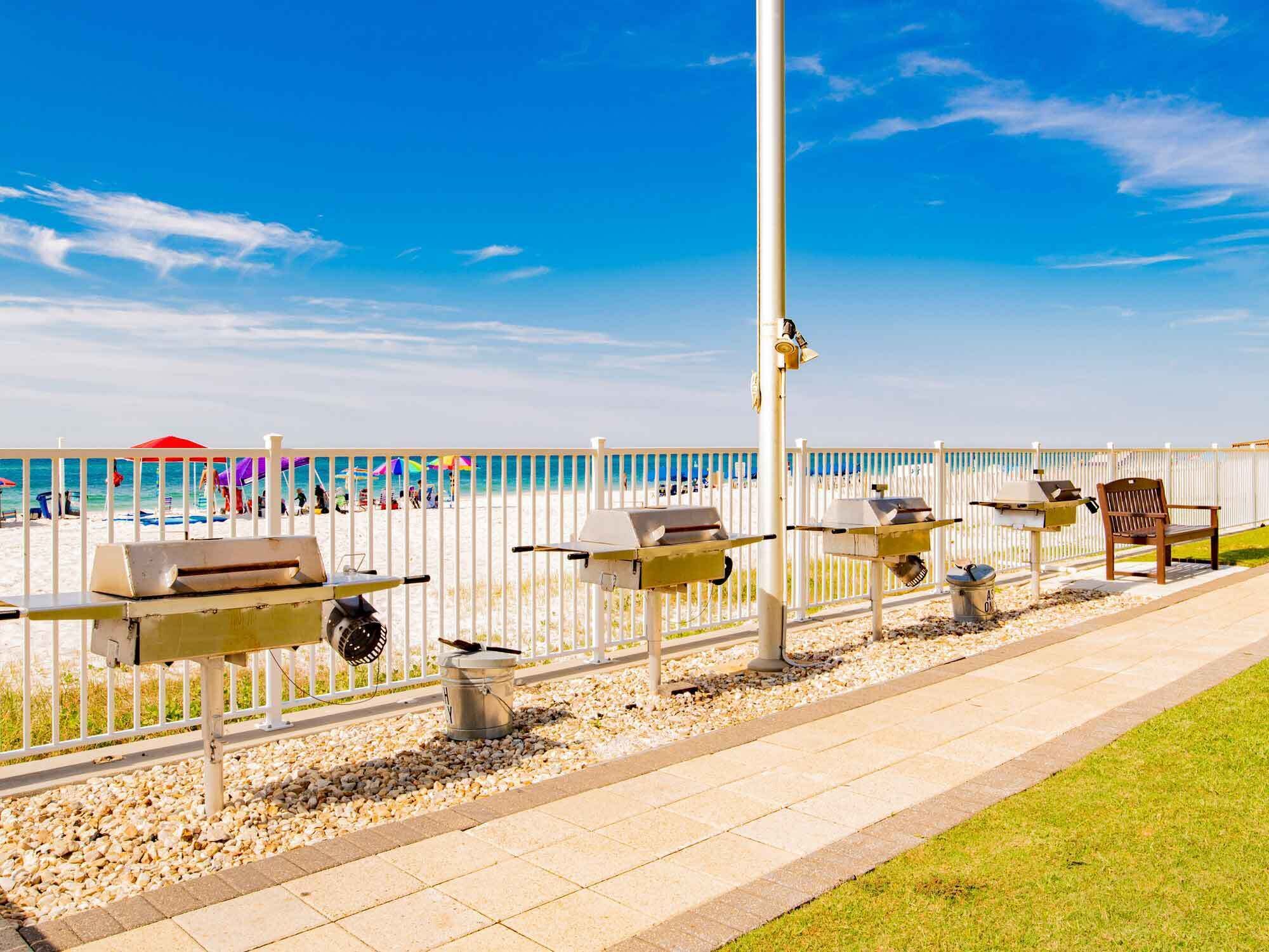 A beachside barbecue area with grills and a grill station, a white fence, and a view of the beach and ocean.
