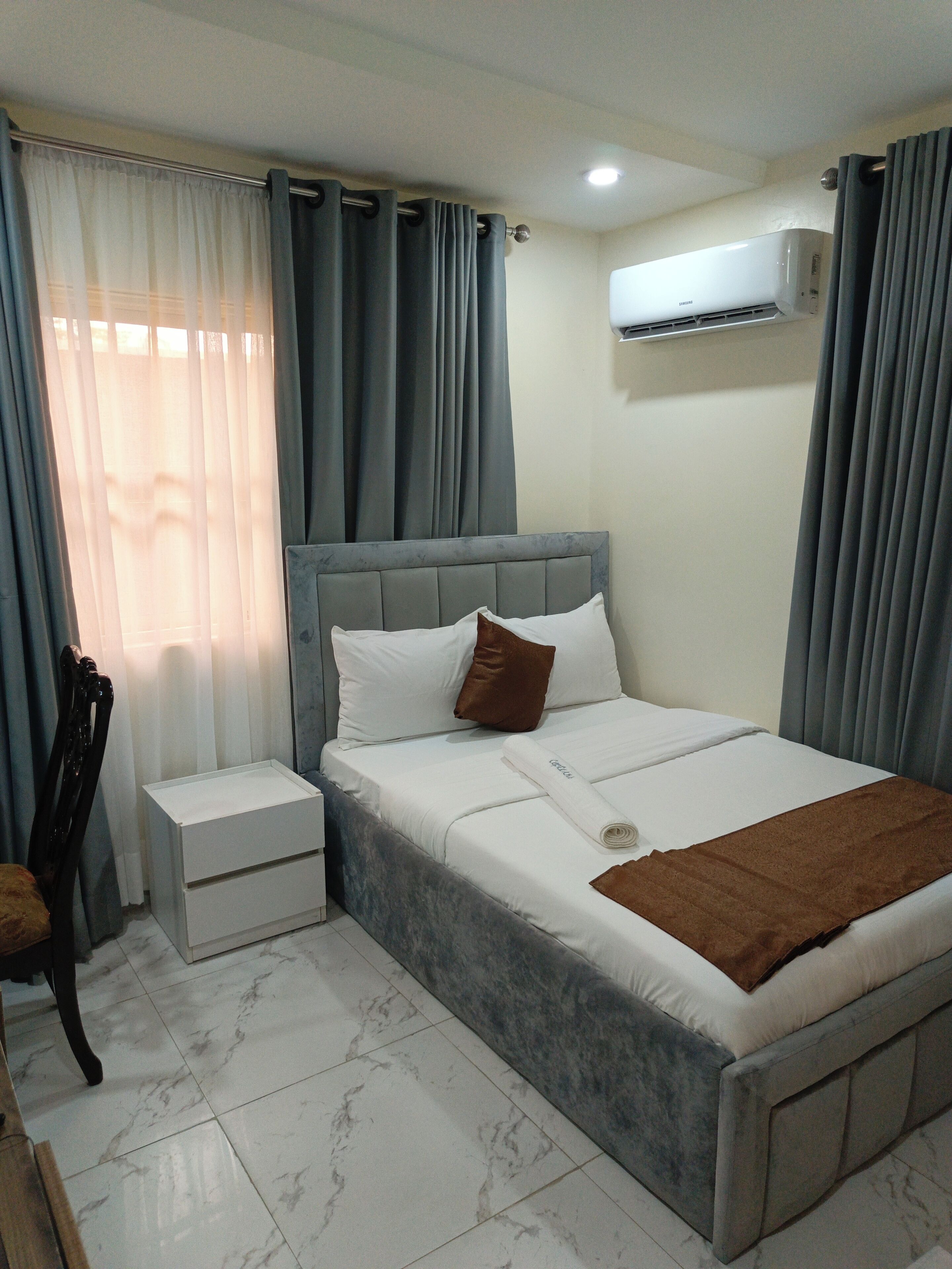 A bedroom with a bed, nightstand, chair, and an air conditioning unit.