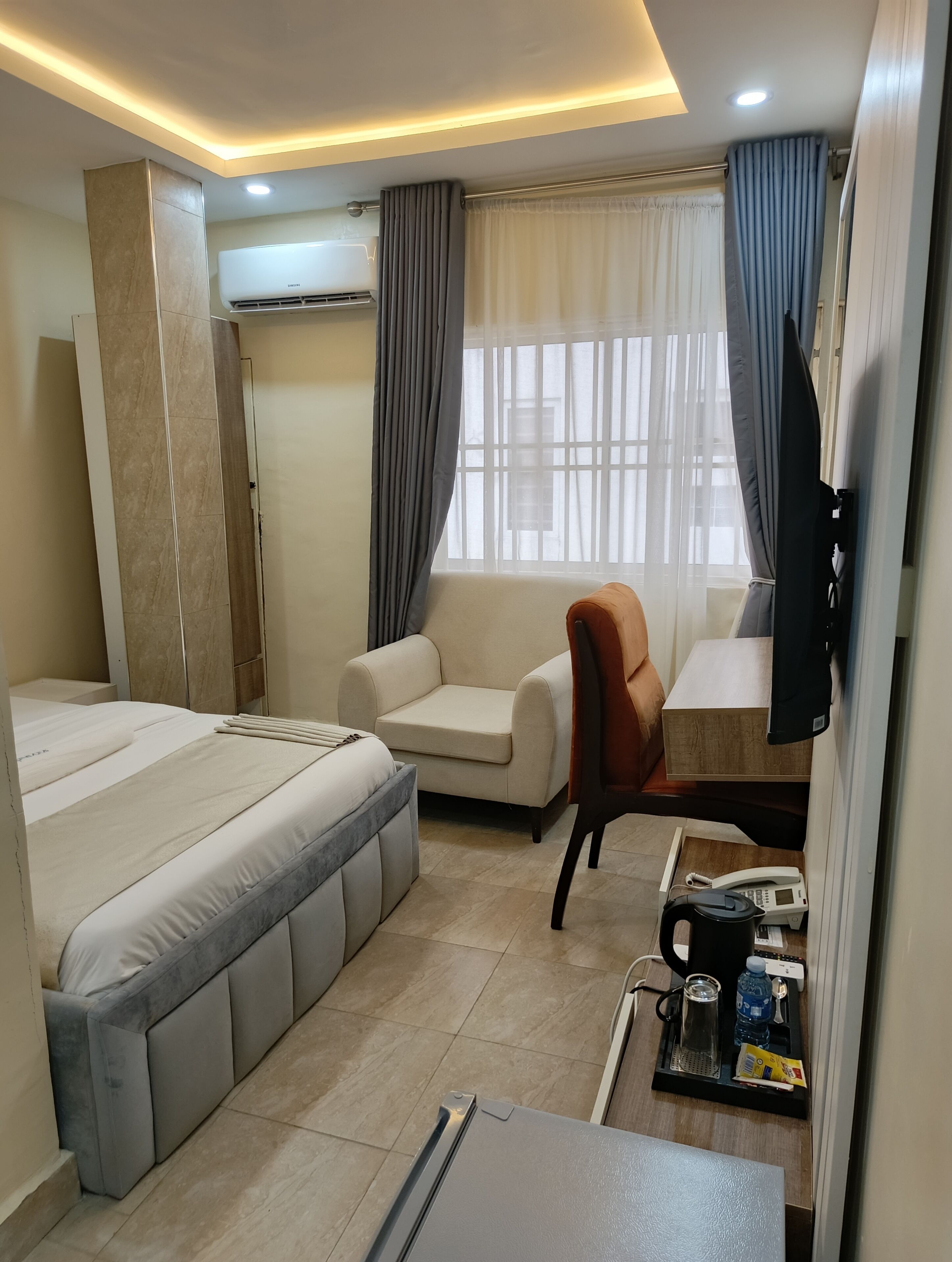 A hotel room with a bed, a sofa, a desk, a TV, and a coffee table with a kettle and water bottle.