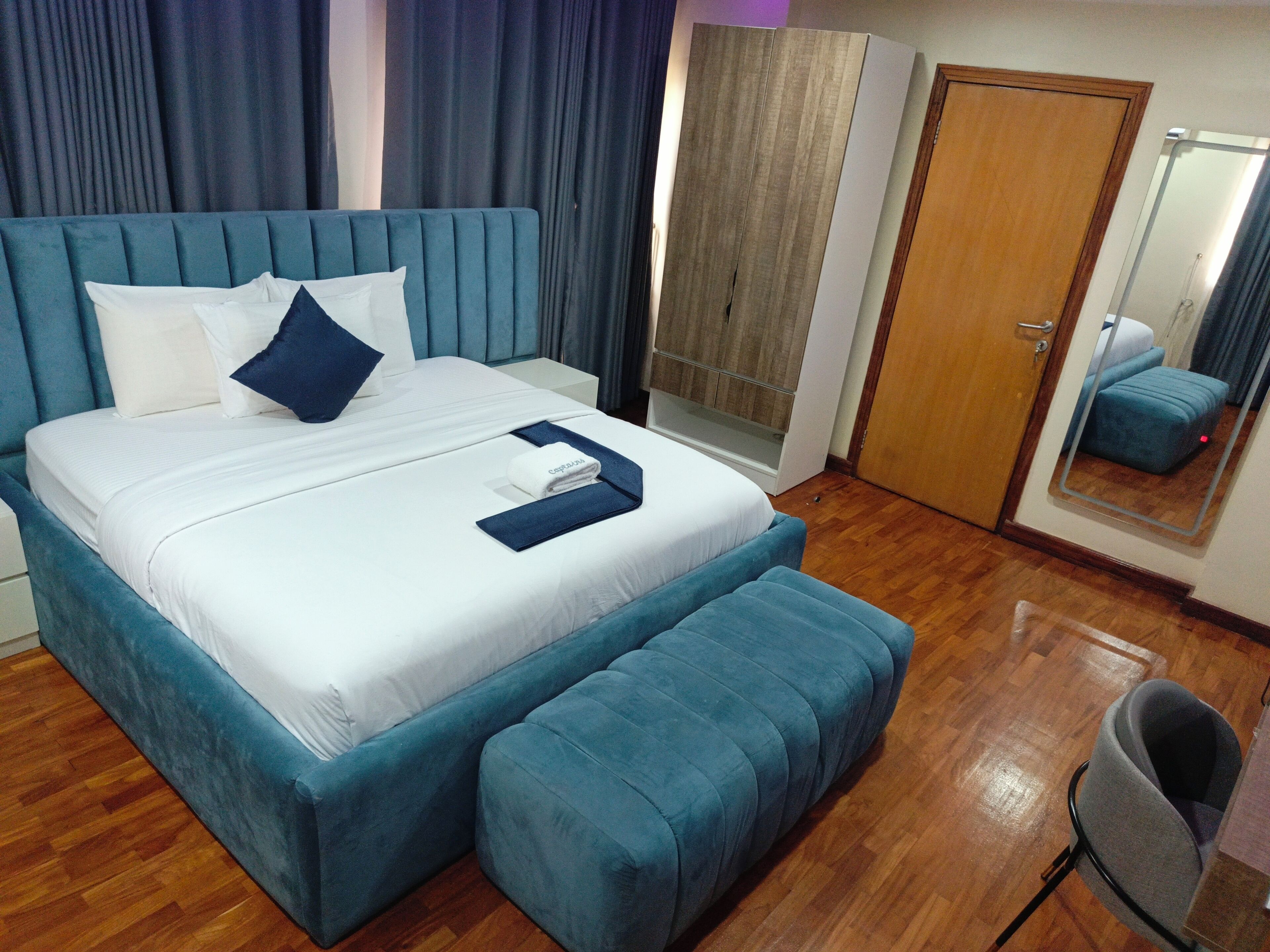A neatly made bed with a blue headboard and pillows, a wooden floor, a small seating area, and a mirror on the wall.