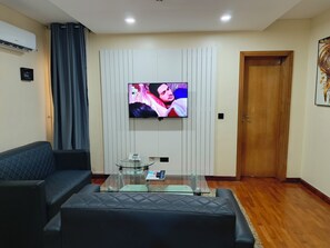 A living room with a flat-screen TV mounted on the wall, a glass coffee table, a black sofa, and a wooden door. - Captain Residency Hotel (Lagos)