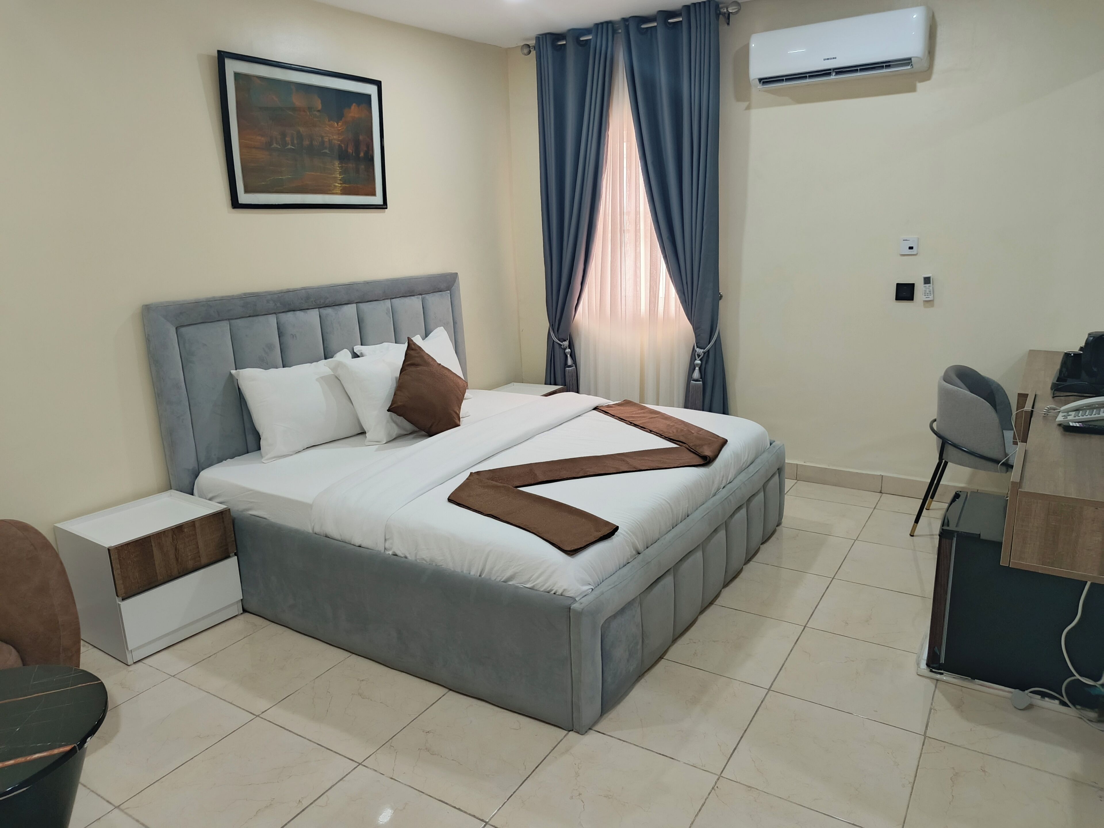 A neatly arranged bedroom with a bed, bedside tables, a desk, and an air conditioning unit.