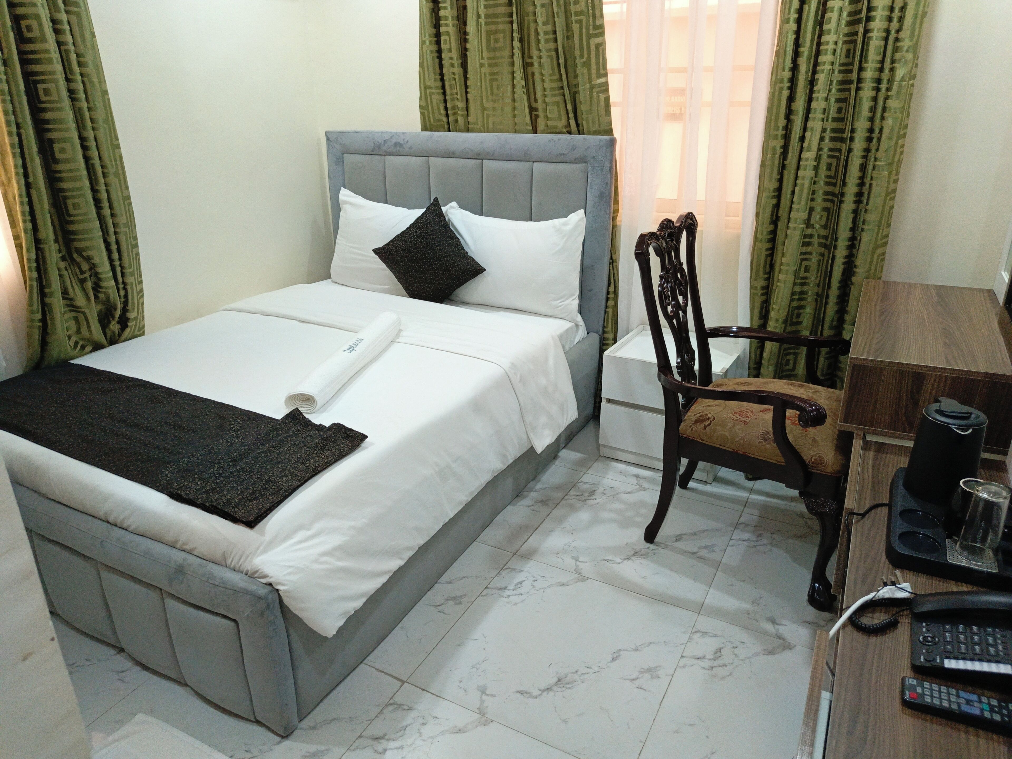 A neatly made bed with white linens and a rolled towel, a wooden chair with a cushion, a desk with a telephone and remote control, and a small white nightstand.