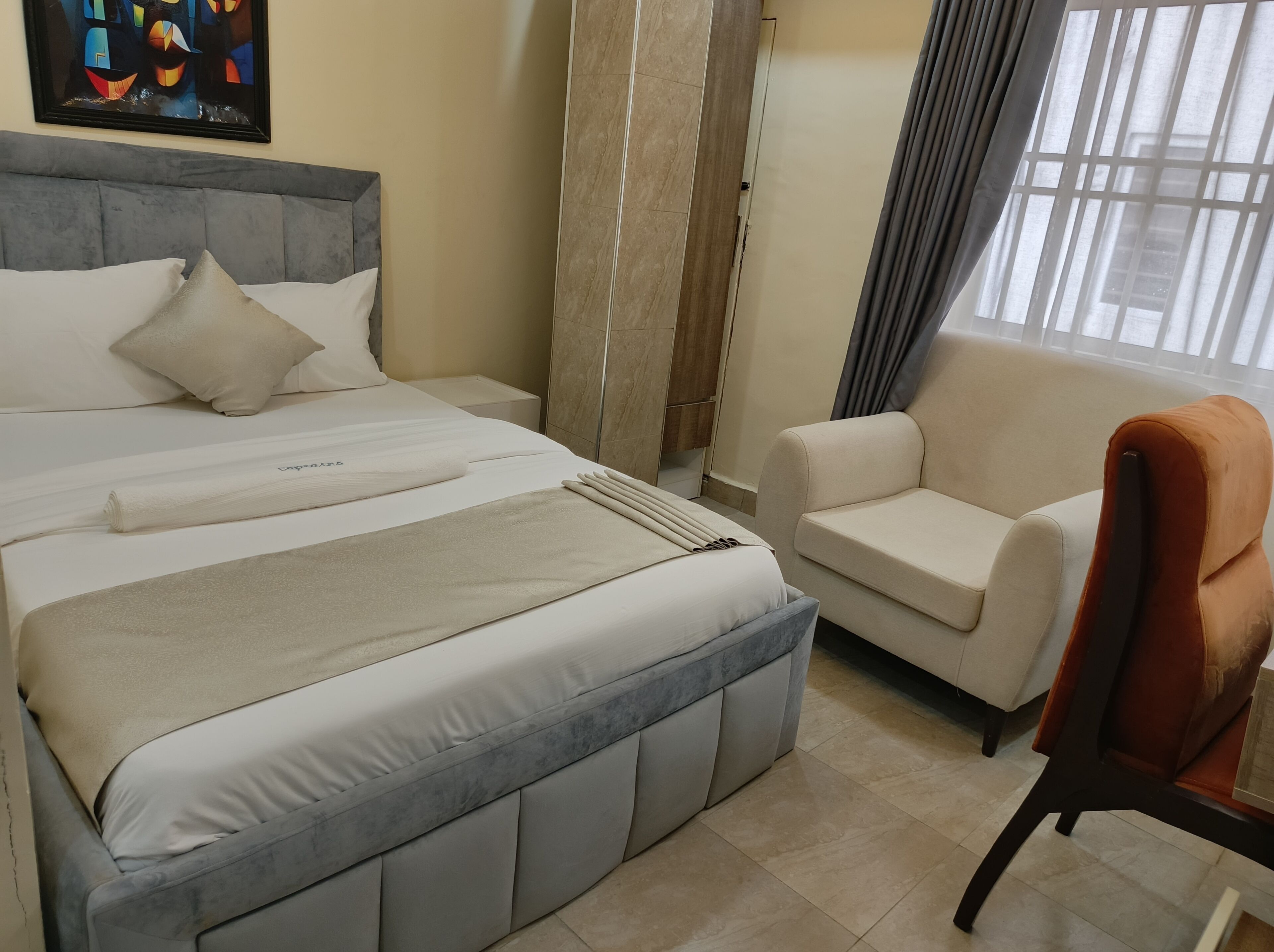 A neatly made bed with a headboard, a bedside table, a chair, and a window with curtains.
