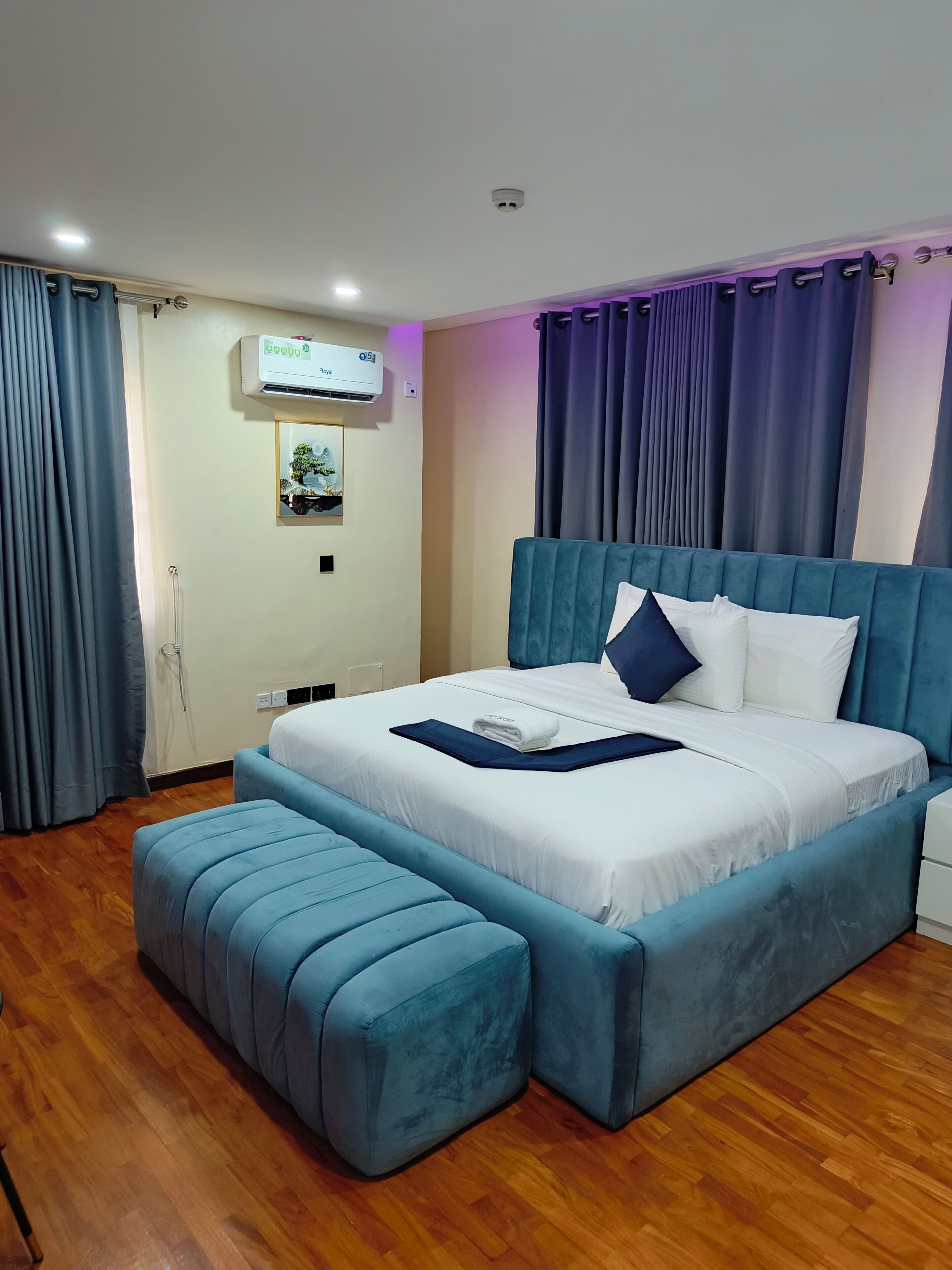 A bedroom with a large bed, a bedside table, a wall-mounted air conditioner, a window with curtains, and a wooden floor.