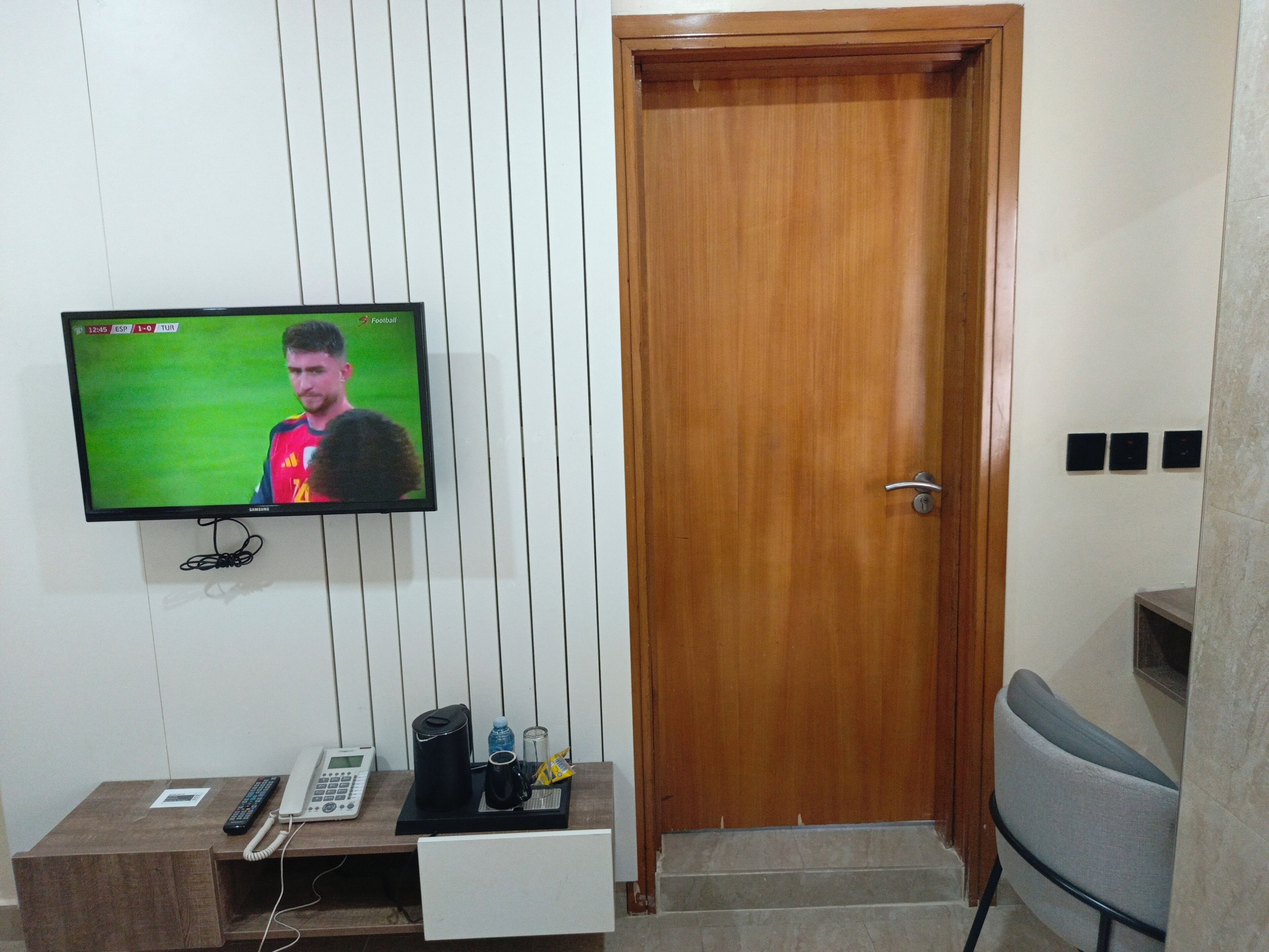 A room with a television mounted on the wall, a wooden door, a desk with a telephone, and a chair.