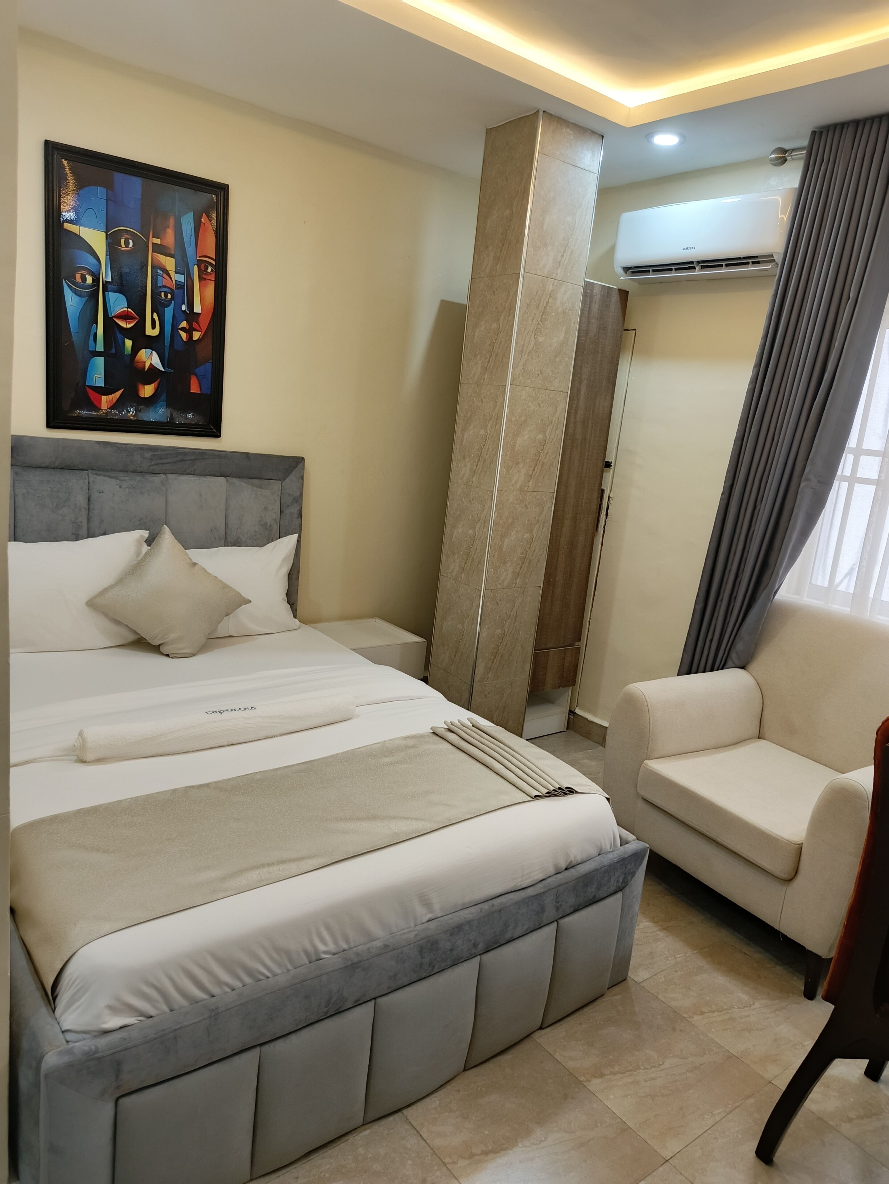 A modern hotel room with a large bed, a sofa, a painting on the wall, and an air conditioning unit.