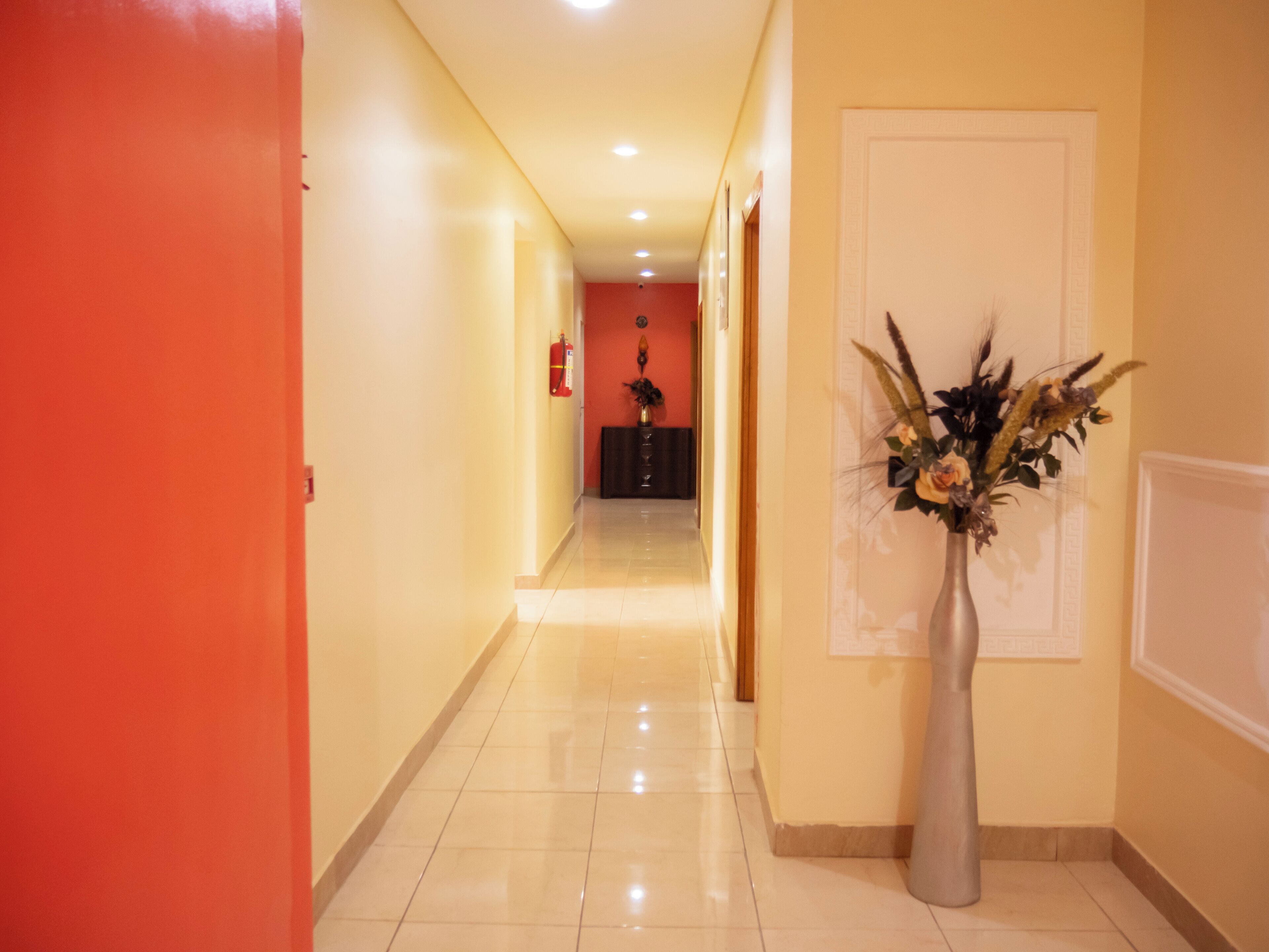 A hallway with a red door on the left, a vase with flowers on the right, and a fire extinguisher on the wall.