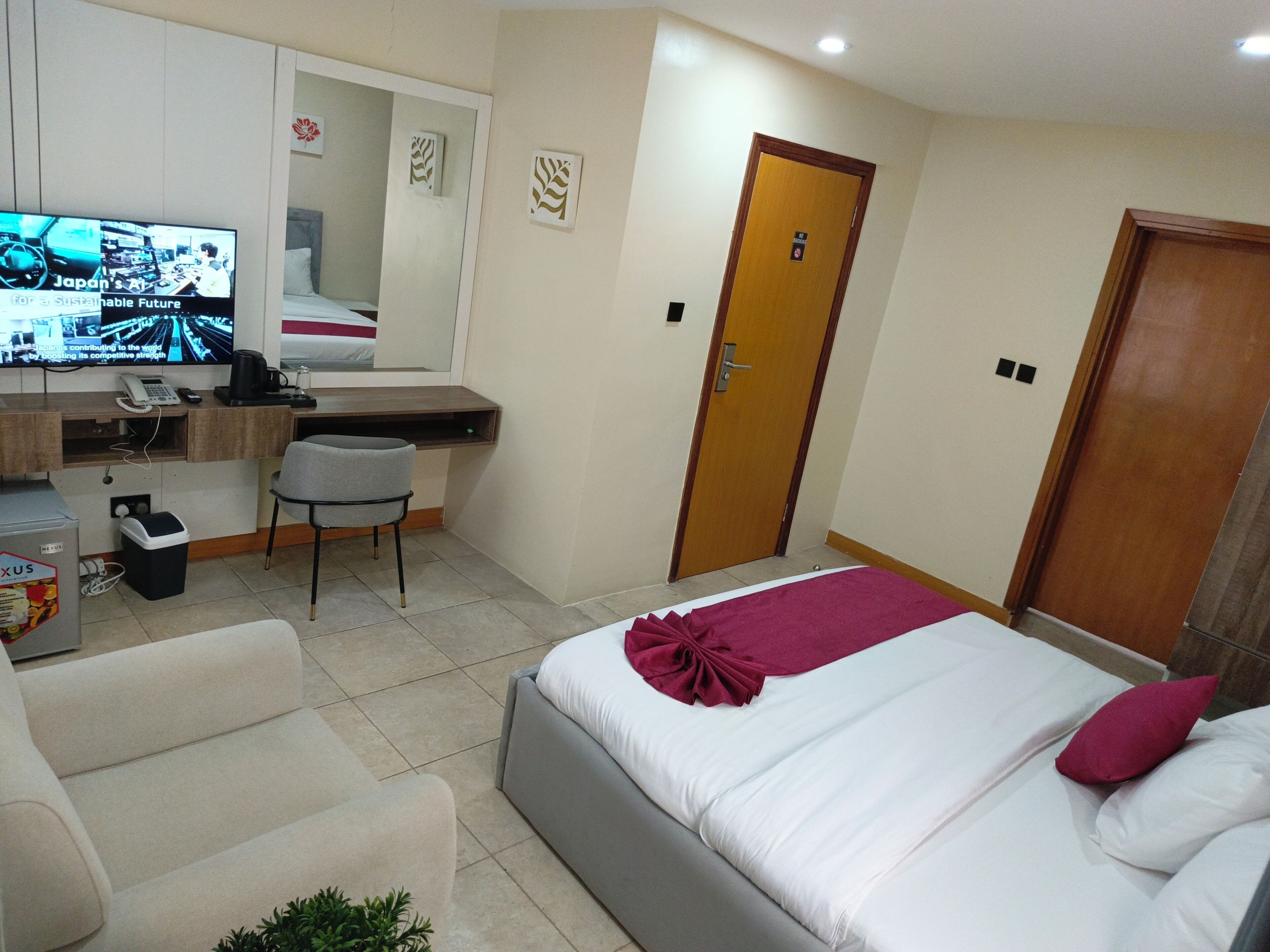 A hotel room with a bed, a desk with a computer, a chair, a trash bin, and a television.