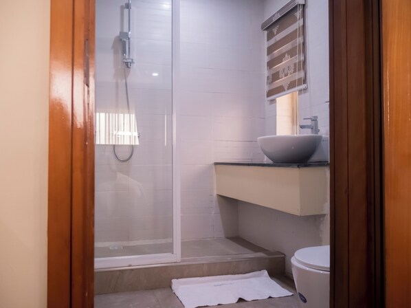 Un baño con cabina de ducha de vidrio, lavamanos blanco con lavabo de superficie y un inodoro.