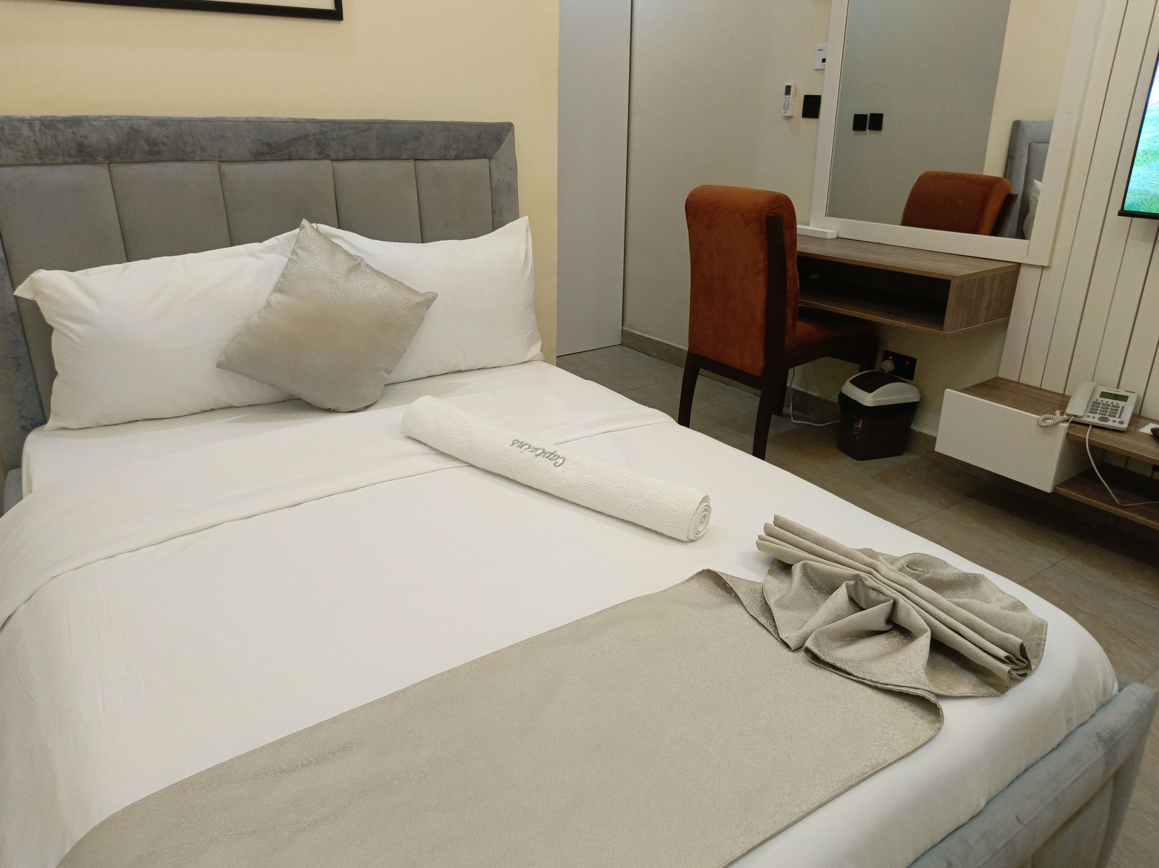 Une chambre d’hôtel avec un lit, des oreillers, une serviette pliée, une chaise, un bureau avec un téléphone et un miroir.