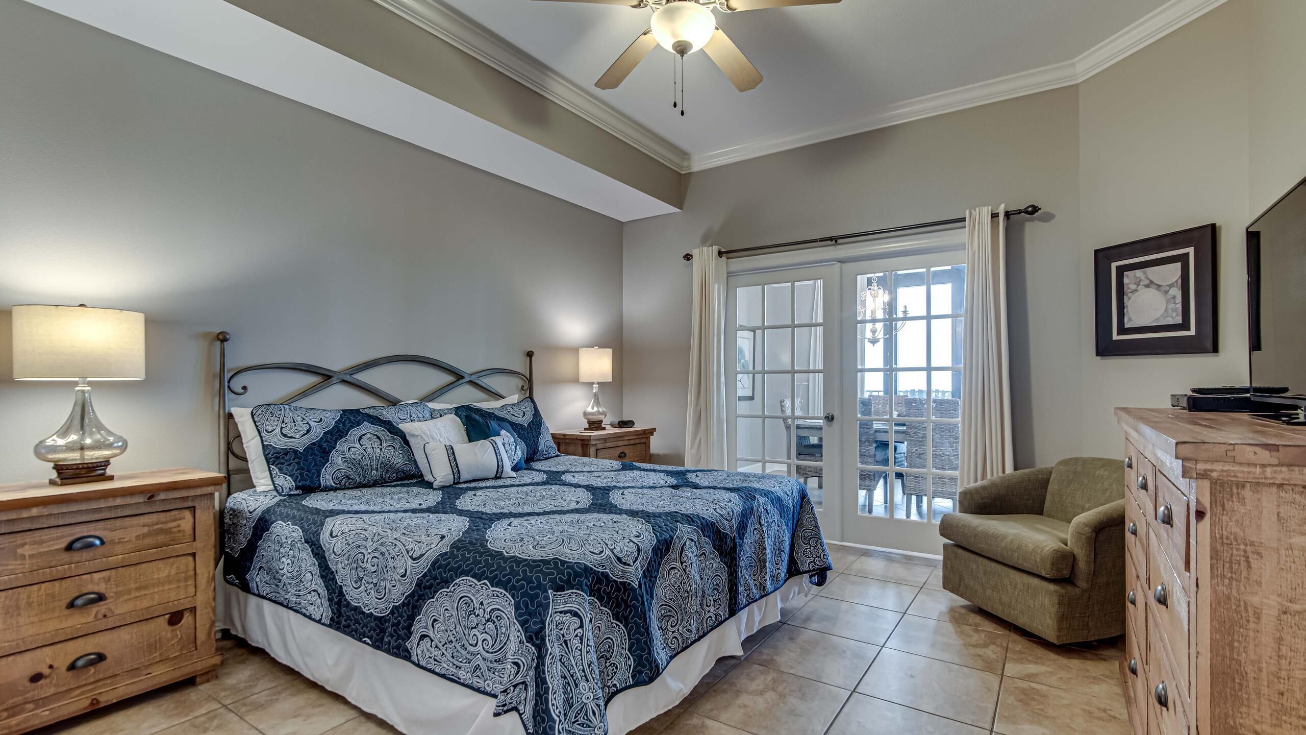 A bedroom with a bed, two bedside tables, a wooden dresser, a chair, and a view through a glass door.