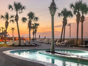 Un espace aménagé au bord de la piscine, avec des chaises longues et des palmiers, offrant une vue sur la plage au coucher du soleil.