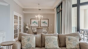 A living room with a beige sofa, floral cushions, a dining table with chairs, a chandelier, and artwork on the wall.