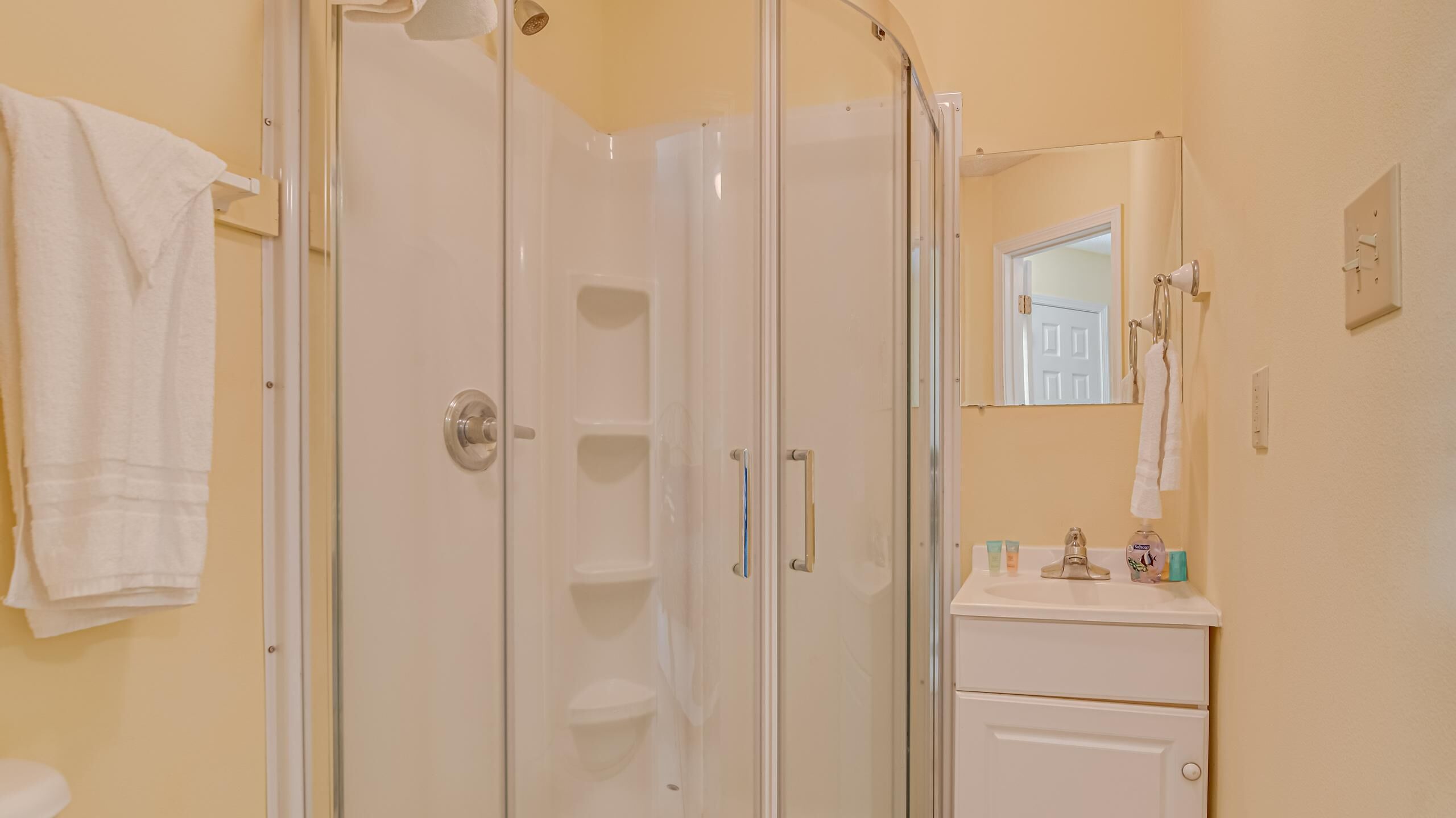A bathroom with a glass shower enclosure, a white sink with toiletries, and a mirror above the sink.