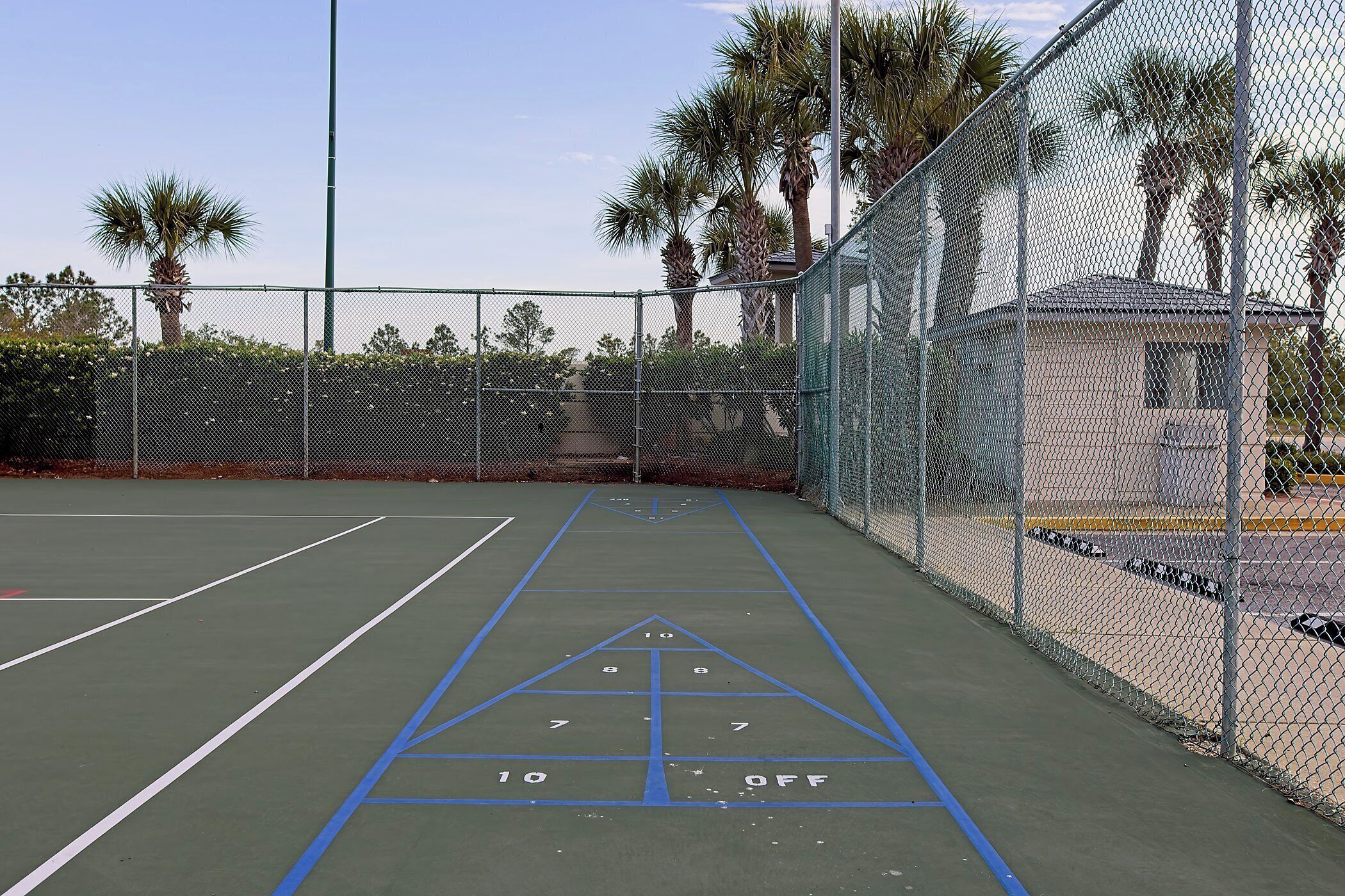A tennis court with a shuffleboard area, surrounded by a chain-link fence and palm trees.