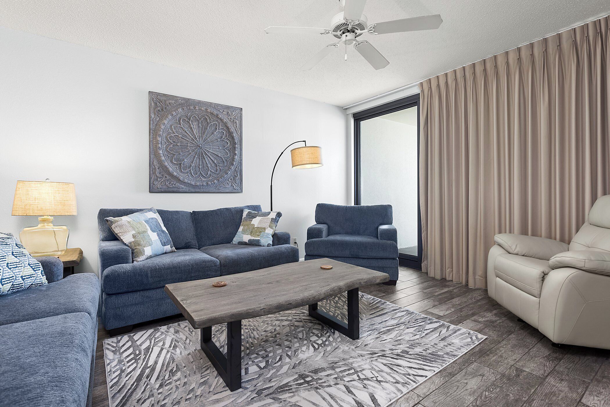 A modern living room with a sofa, armchair, and coffee table, a ceiling fan, and a large window with curtains.
