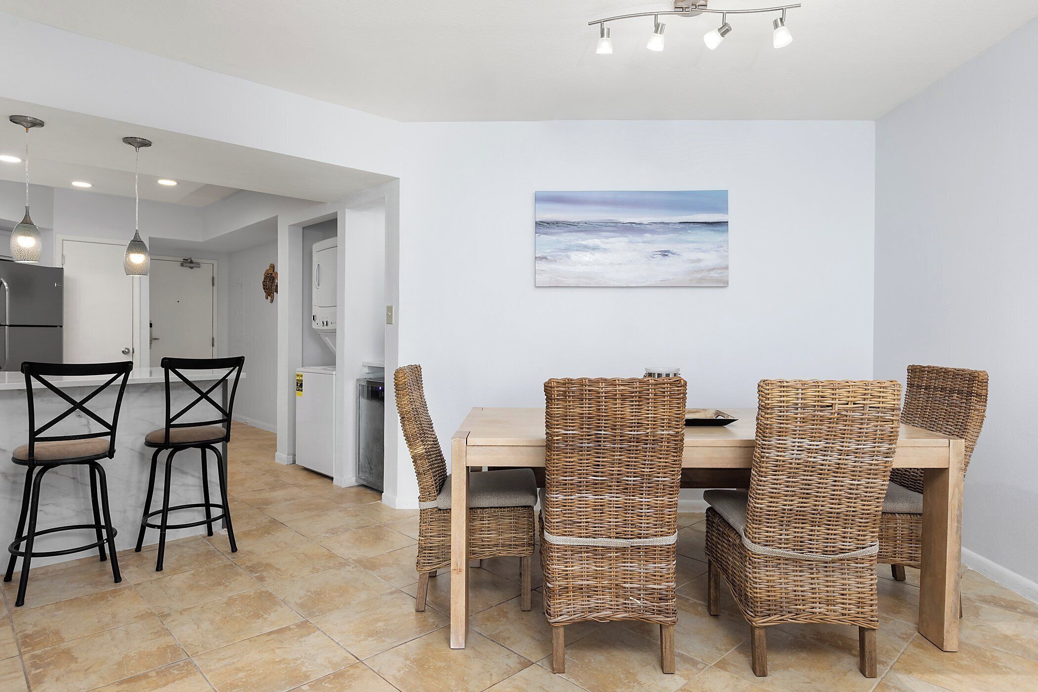 Ein Essbereich mit einem Holztisch und Korbstühlen, einem Strandbild an der Wand und einer Küche im Hintergrund.