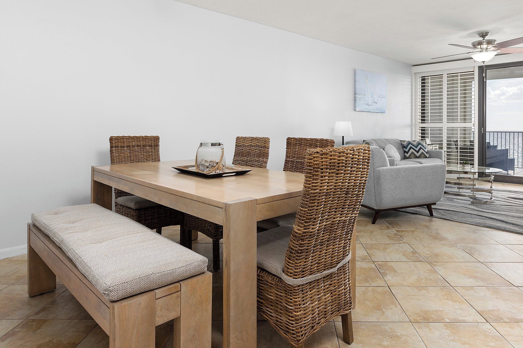 Un coin repas avec une table en bois, des chaises en osier et un banc. Il y a un balcon avec vue sur l’eau.