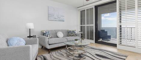 Ein modernes Wohnzimmer mit einem grauen Sofa, einem Glastisch, einem Deckenventilator und Blick auf den Balkon.