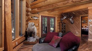 A log cabin interior with a bear and deer mounted on the walls, a leather couch with red cushions, and a wooden floor. - Grand Wildlife Lodge 10ppl (La Pine)