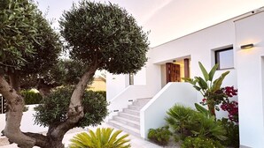 Una casa moderna amb façana blanca, una escala que condueix a l'entrada i diverses plantes i arbres al jardí.
