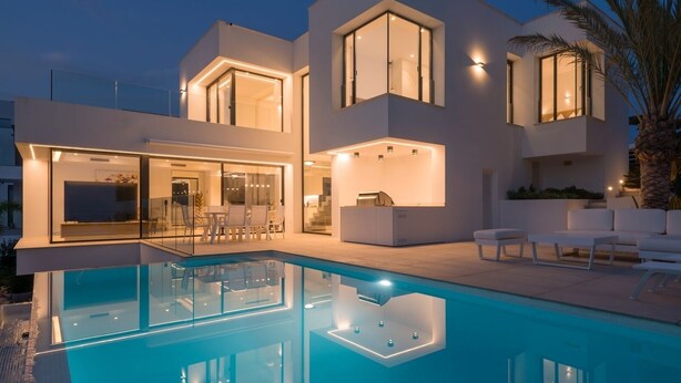 Una casa moderna de diversos pisos amb piscina, grans finestres i mobiliari d'exterior.