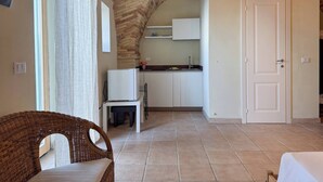 Una habitación con una mesa de comedor, una silla de mimbre, una zona de cocina con armarios blancos y una puerta.