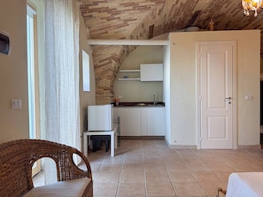 A room with a dining table, a wicker chair, a kitchen area with white cabinets, and a door.