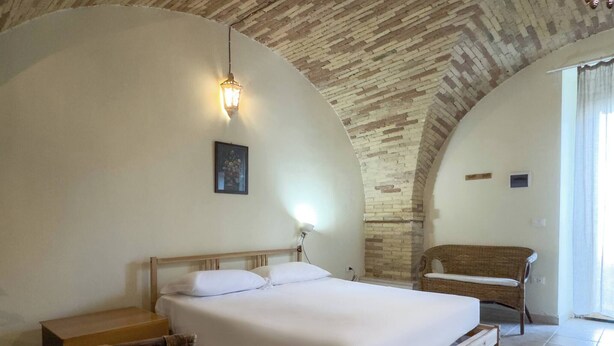 Un dormitorio con cama, mesita de noche, una silla, un banco de madera, una lámpara de pared y techo de ladrillo.