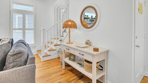 Un escalier avec des marches en bois et des rampes blanches, une console blanche surmontée d’une lampe, des paniers en osier et un tableau encadré.