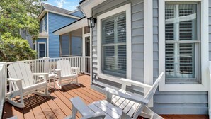 Une terrasse en bois avec des fauteuils Adirondack blancs, une balustrade blanche et une grande fenêtre avec des volets.