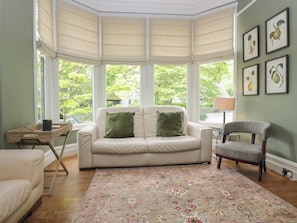 A room with a bay window, a white sofa with green cushions, a patterned rug, and framed botanical prints on the wall.