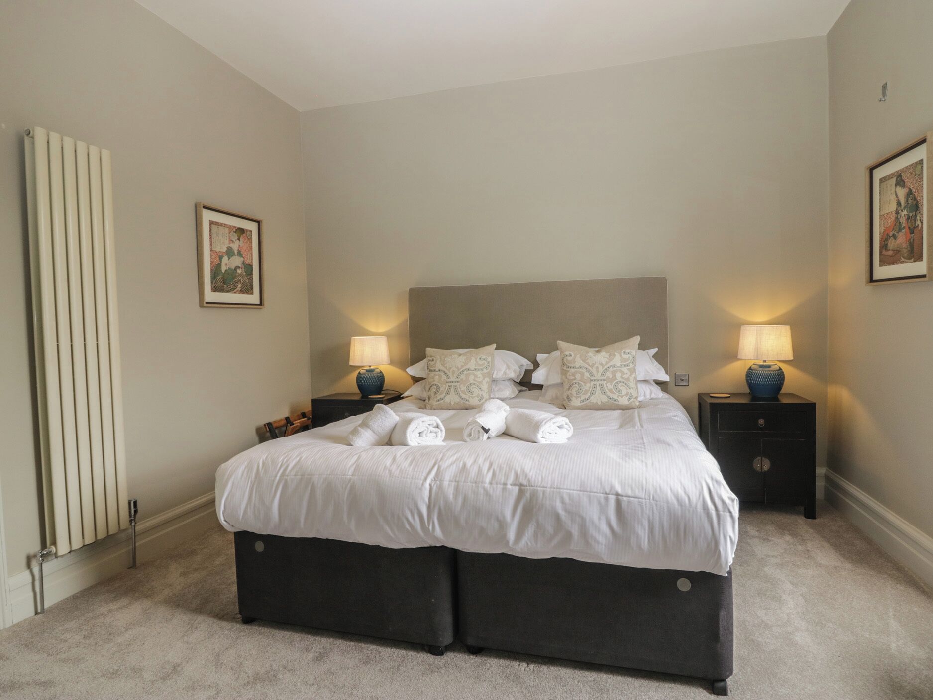 Une chambre à coucher bien rangée, avec un grand lit, deux tables de chevet avec des lampes, un radiateur et des œuvres d’art encadrées accrochées aux murs.