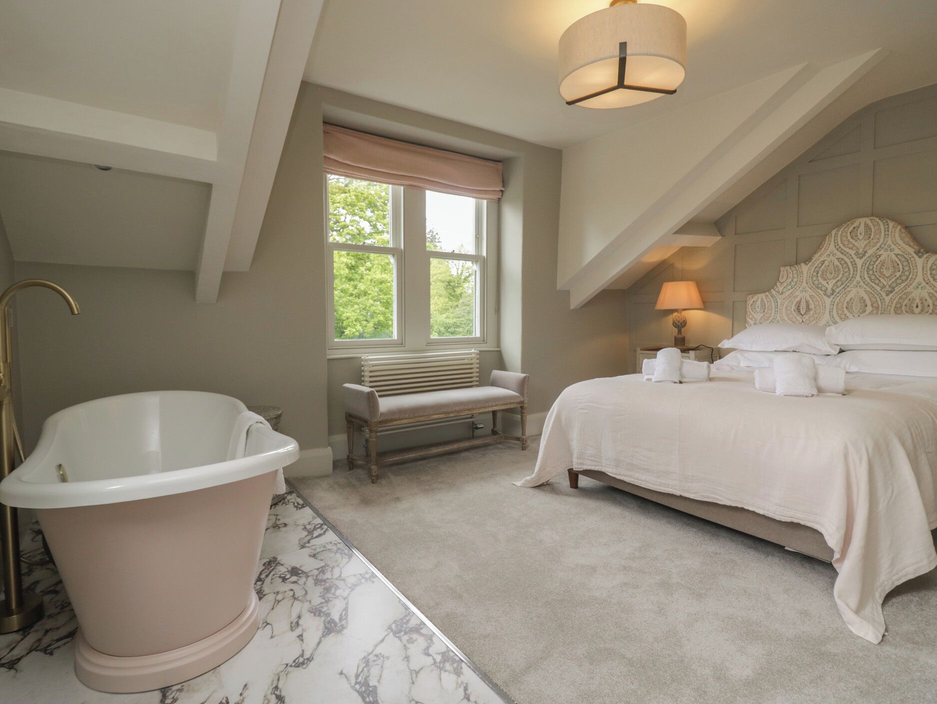 Une chambre avec une grande baignoire, un banc, un lit avec du linge de lit blanc, une fenêtre donnant sur la verdure et un luminaire au plafond.