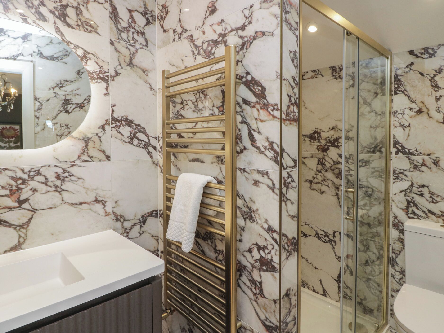 Un baño con pared de mármol, toallero dorado, cabina de ducha de vidrio y lavamanos blanco.
