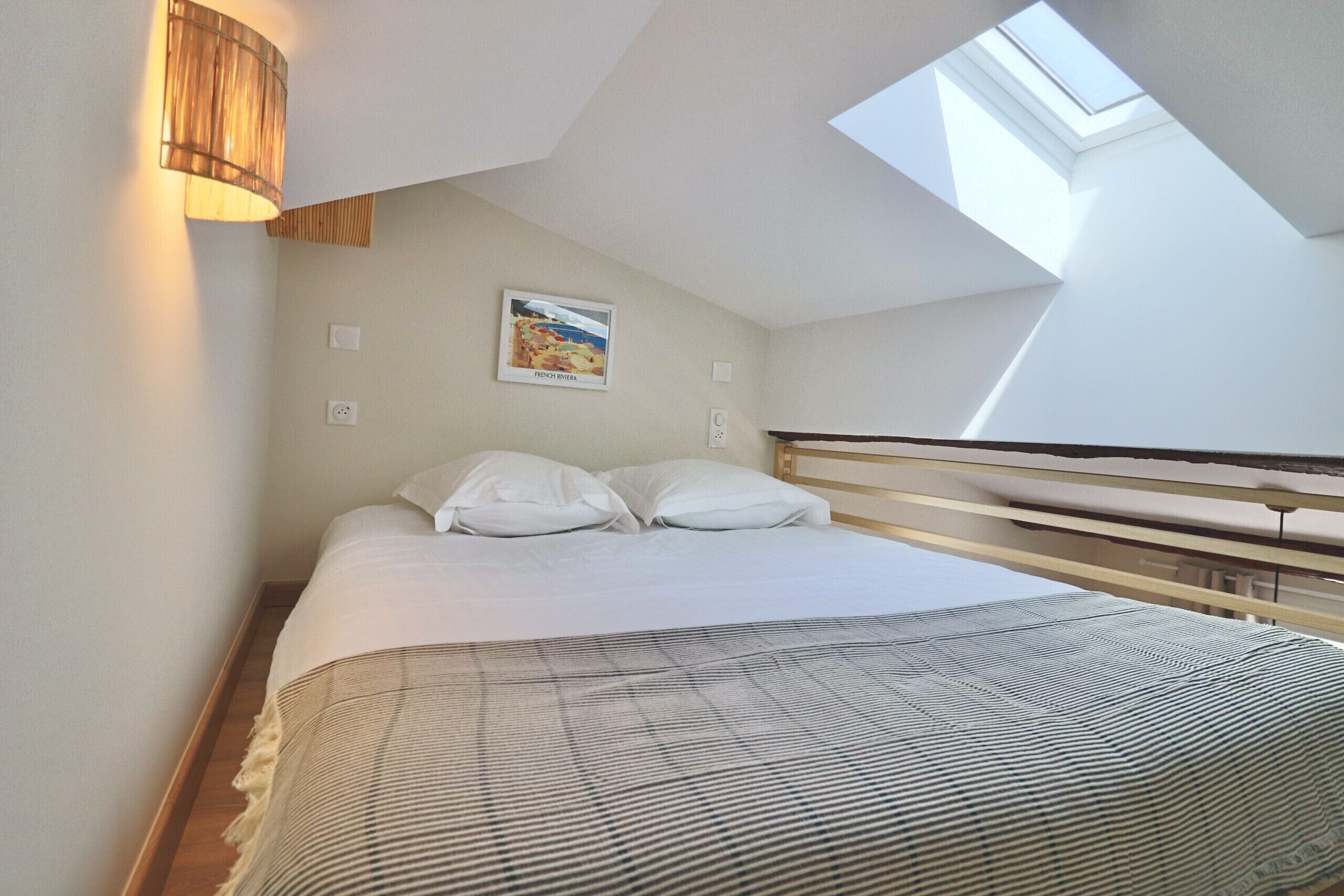 Un dormitorio con tragaluz, cama con ropa blanca, lámpara de pared y una pintura de una escena de playa.