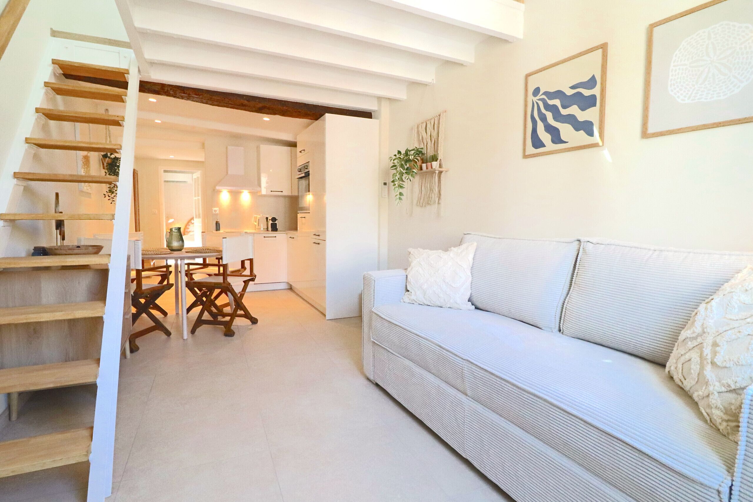 A modern living room with a white sofa, wooden staircase, and a kitchen in the background.