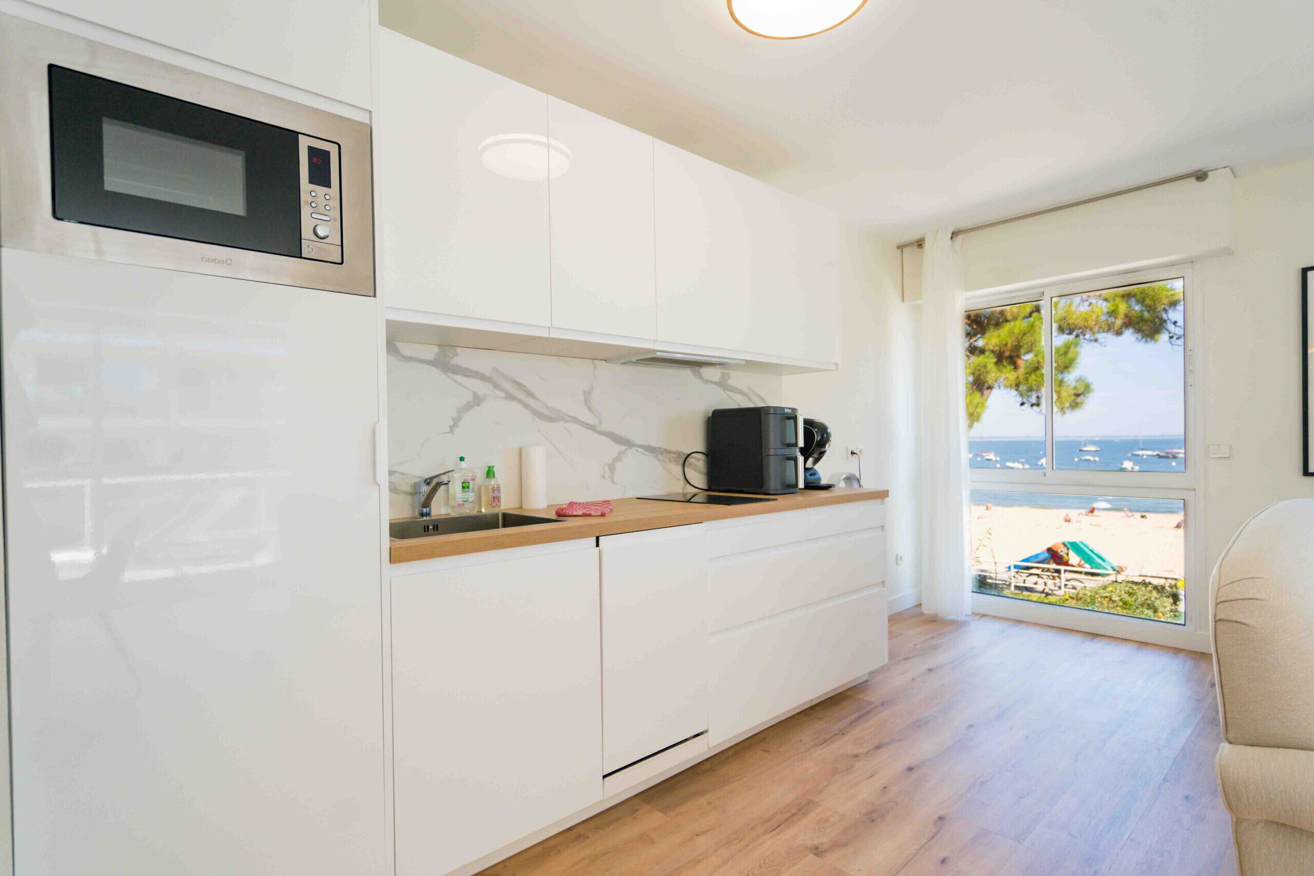 A modern kitchen with white cabinets, a built-in microwave, and a coffee machine. There is a window with a view of the sea and a beach.