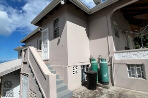 A two-story building with a staircase leading to the entrance, a garage door, and a green gas cylinder. - Ocean View 3-bedroom Apartment in Grand Anse with AC (Saint George)