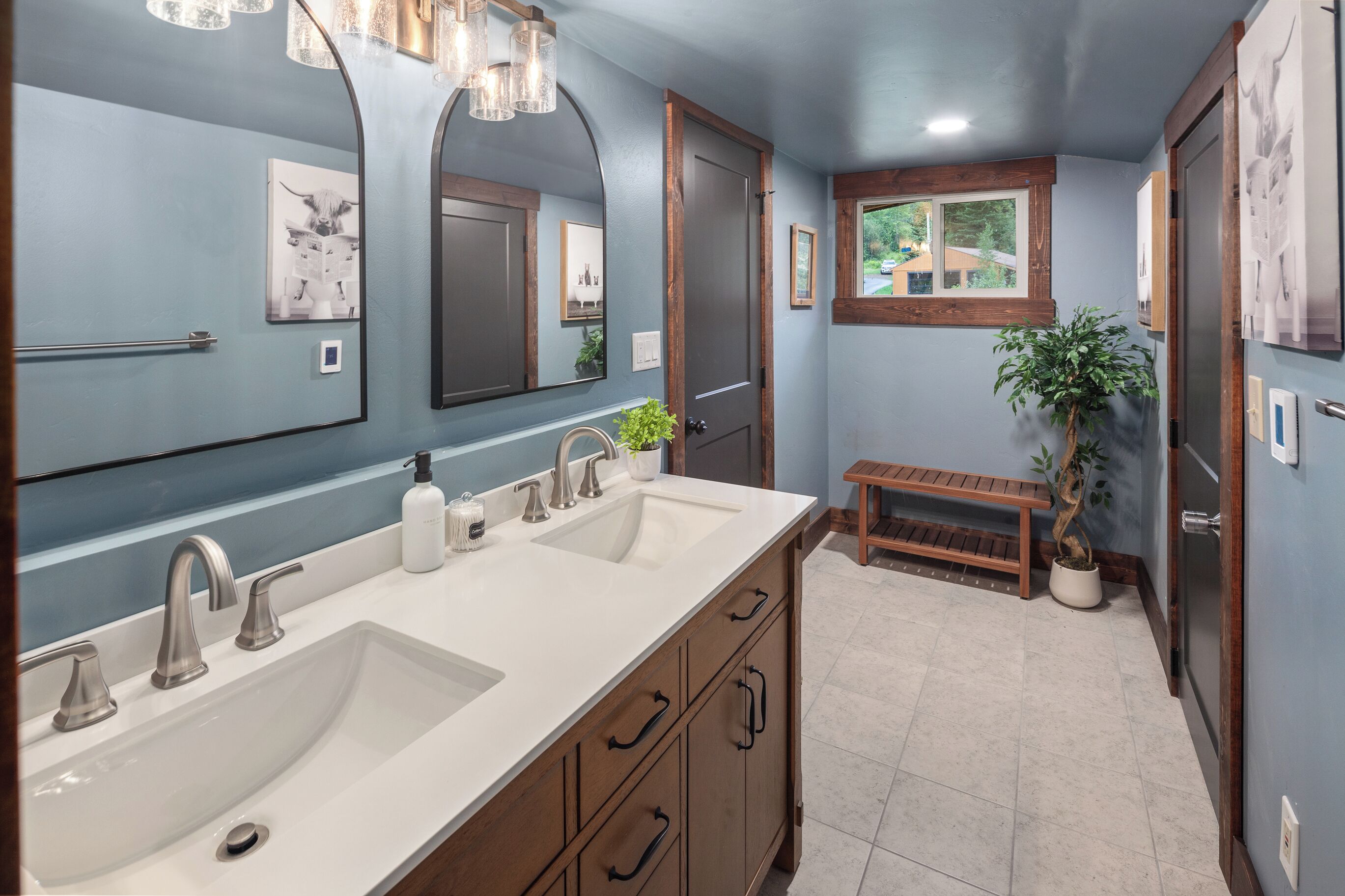 A bathroom with a double sink, wooden cabinets, a bench, and a potted plant.