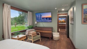 Un dormitorio con una cama grande, una silla, un televisor montado en la pared, una ventana con cortinas y vistas al exterior.